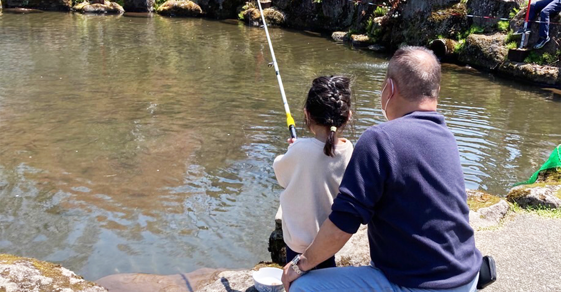 ホテル若月ファミリー釣り堀 — 長野県信濃町・黒姫高原 · 大人が後ろに付き添い、子どもが池に竿を出すえさ釣りの様子