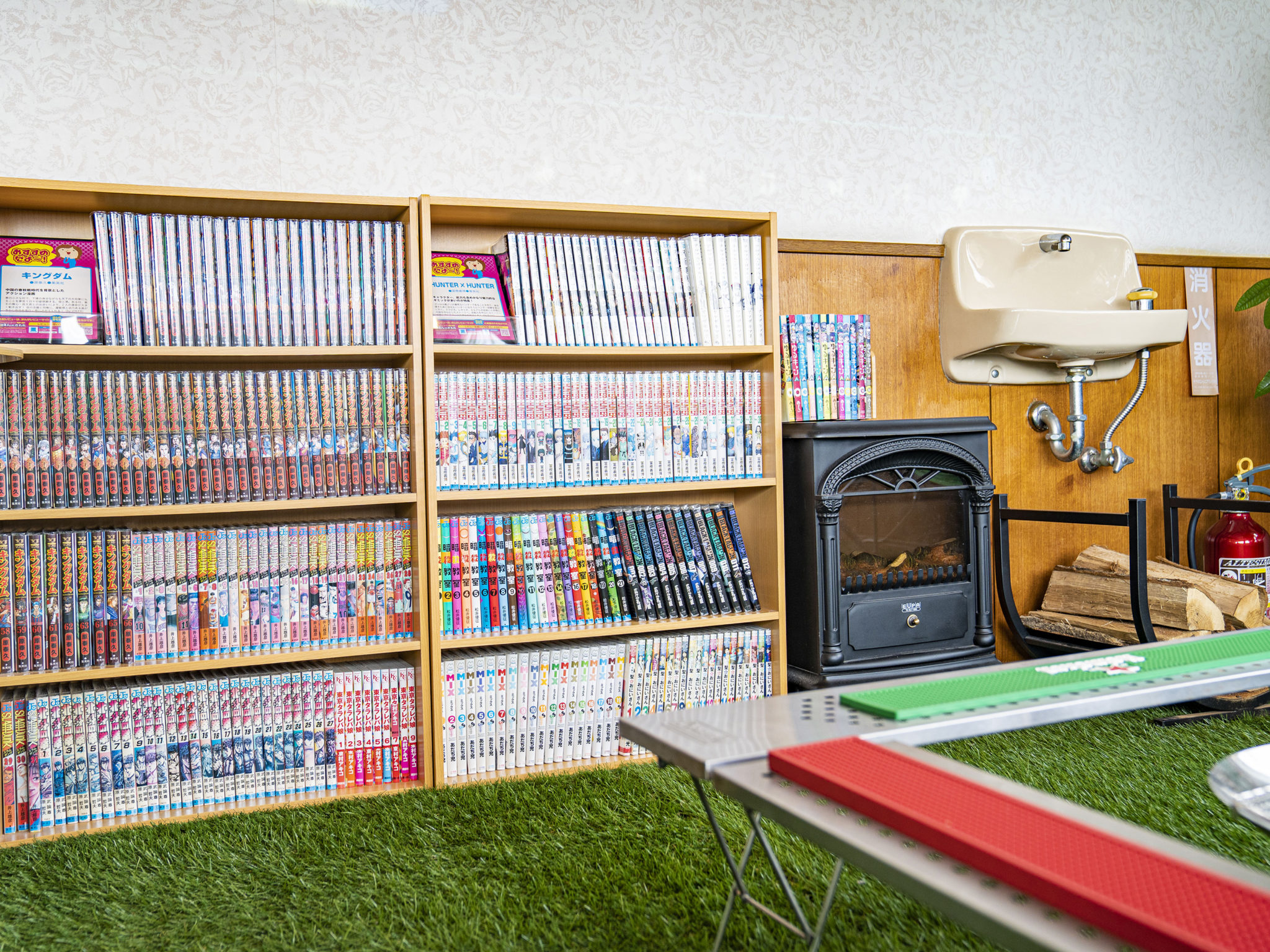 We Home Villa 城ケ崎温泉 — 漫画が並ぶ棚と人工芝のパターゴルフスペース、暖炉型ヒーターのある共用部