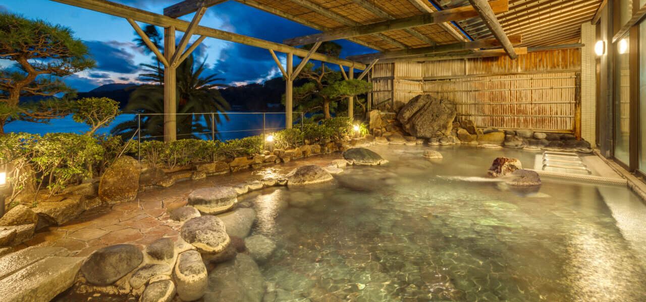 稲取銀水荘 — 静岡・稲取温泉 · 夕暮れの海を望む岩造りの展望露天風呂、脱衣所にベビーベッドとベビーバスを備える